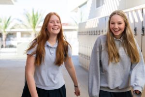 Two girls are laughing, walking down a sunlit hallway of lockers.